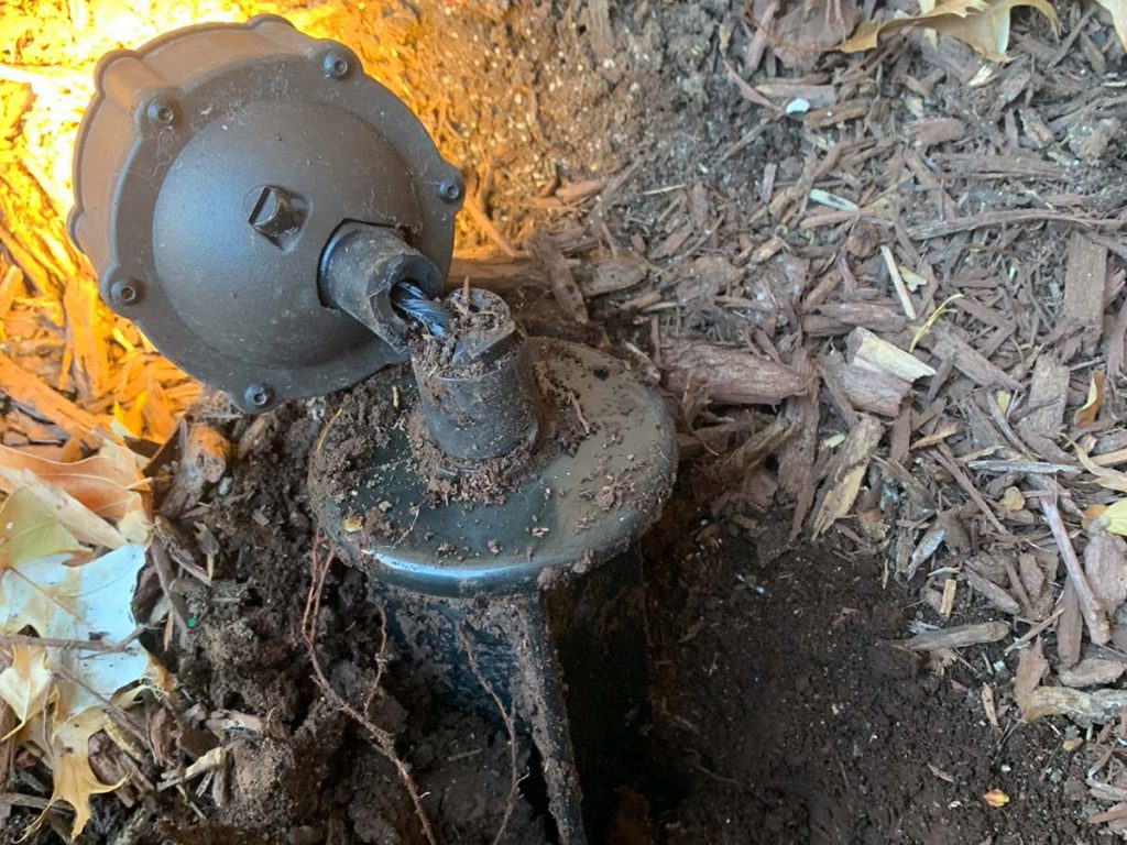 A close-up view shows a landscape light fixture with its top housing detached, revealing the internal wiring connected to a ground stake. The unit is partially buried in dark soil and wood mulch.