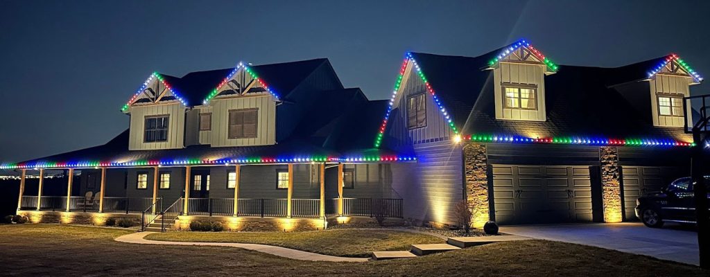 The lighting design provides clear visibility of the surrounding lawn, stone walkways, and the multi-car garage.
