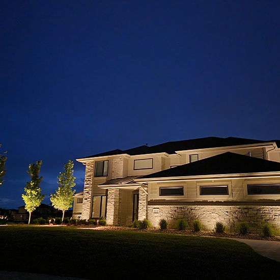 This modern home features a light stone and white siding facade illuminated by warm wall sconces. Bright ground-mounted spotlights highlight three tall, slender trees in the front yard, casting vibrant green textures against the night sky.