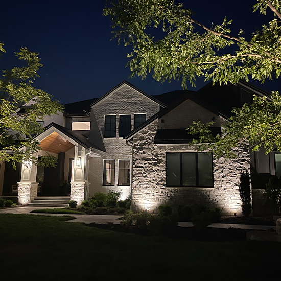 This modern home features a light stone and white siding facade illuminated by warm wall sconces. Bright ground-mounted spotlights highlight three tall, slender trees in the front yard, casting vibrant green textures against the dark night sky.