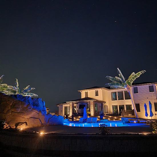 Warm accent lights illuminate the modern house's siding while blue underwater LEDs glow within the pool. Integrated step lights line the patio stairs, and a lighted palm tree sculpture stands near the water's edge.