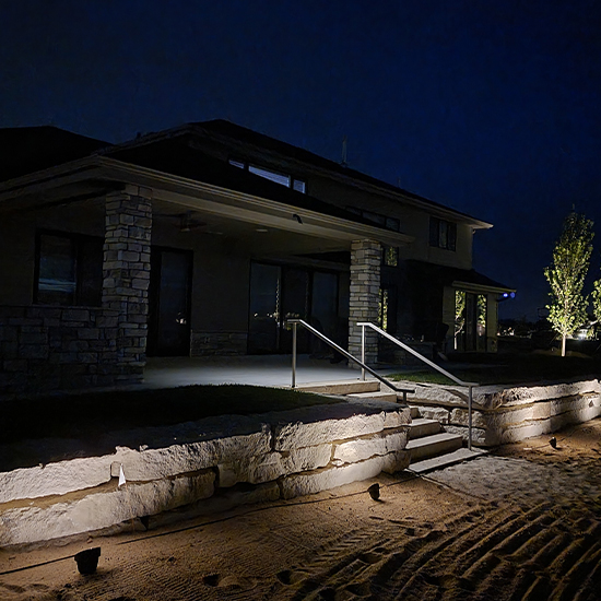This tiered stone patio features a dark siding house with multiple levels. Numerous recessed LED step lights are integrated into the masonry stairs, providing a warm, guiding glow across the entire exterior landscape and walls.