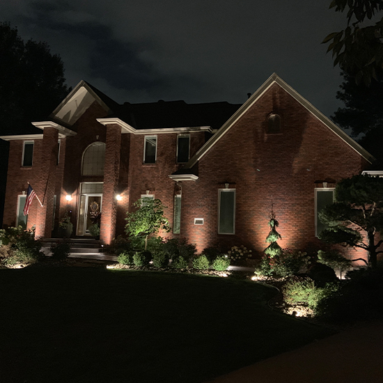 Two-story red brick home features a grand arched entryway illuminated by symmetrical wall sconces. Warm landscape spotlights highlight the facade and manicured shrubs, while a small American flag hangs near the front porch.