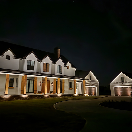 This modern farmhouse features bright white siding with large wood-timber columns supporting a wide front porch. Warm landscape spotlights illuminate the foundation, while recessed gabled lighting emphasizes the roofline and detached garage.