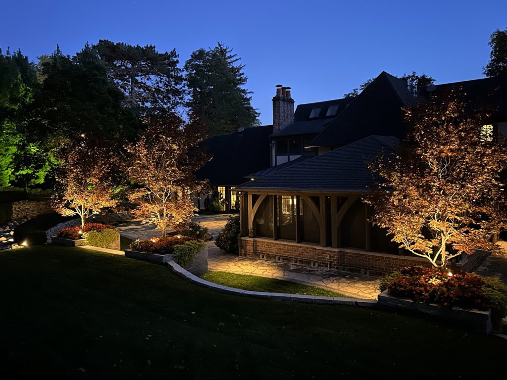 A multi-level backyard features stone pathways and planters illuminated by warm landscape spotlights. These lights highlight several Japanese maples and garden beds, while the adjacent brick and timber house is lit by warm interior window glows.