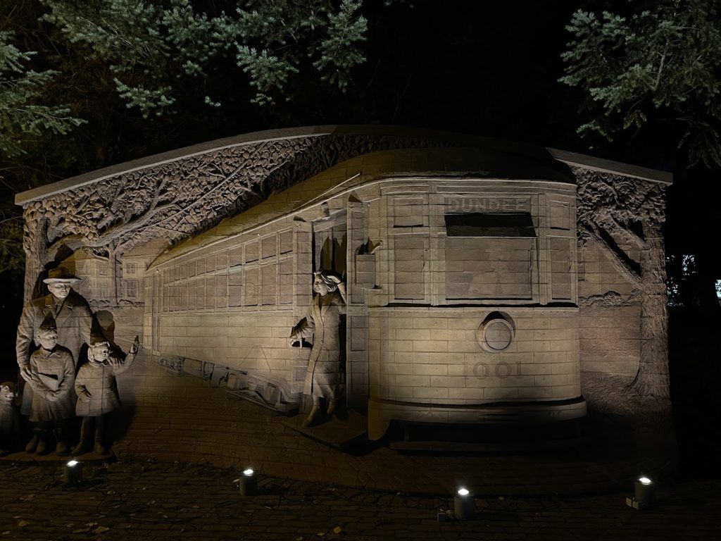 Large, detailed stone relief sculpture depicts a historical scene featuring a "Dundee" trolley car and figures in mid-century attire. Multiple ground-mounted spotlights are positioned along the base, casting a warm, even glow that highlights the intricate textures of the stone carvings and the surrounding evergreen branches.