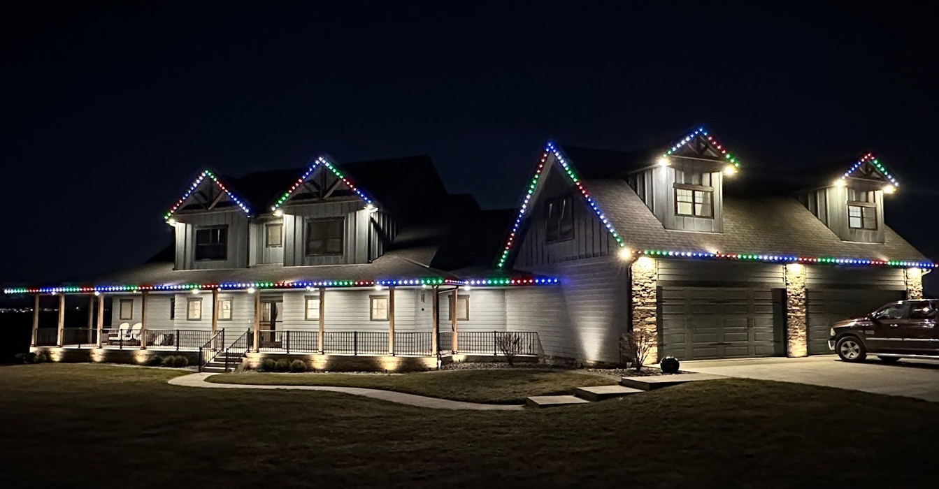 Large craftsman-style house features a wide front porch and multiple gables outlined with vibrant, multi-colored holiday lights. Warm spotlights illuminate the stone pillars and garage area, providing a sophisticated architectural contrast.