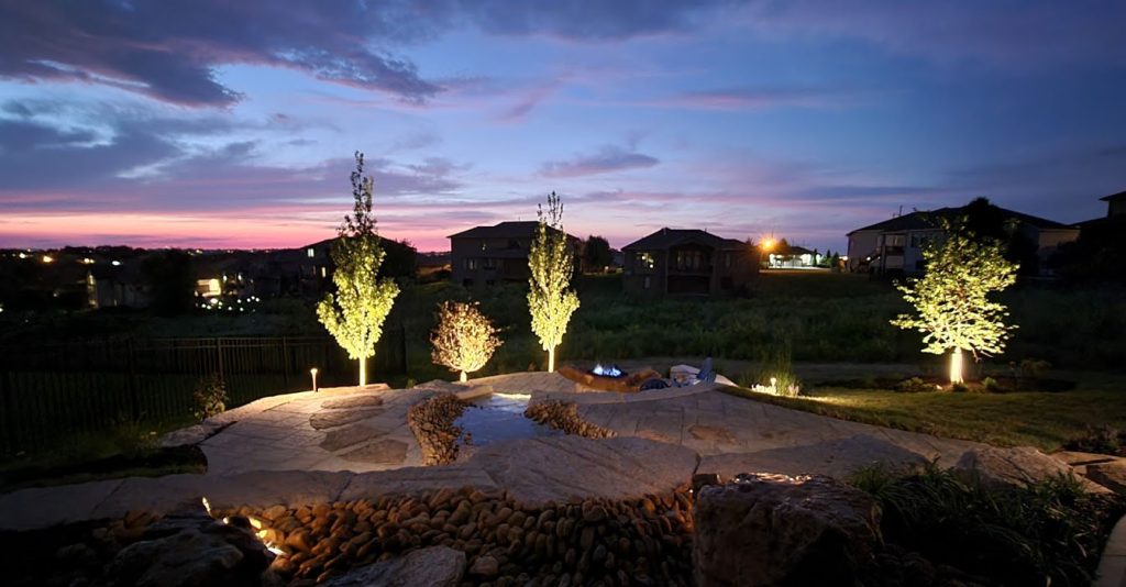 Stone patio and water feature are illuminated by warm landscape lighting at sunset. Young trees feature bright uplighting, while integrated path lights and accent lights highlight the textured stone surfaces and cascading rocks.