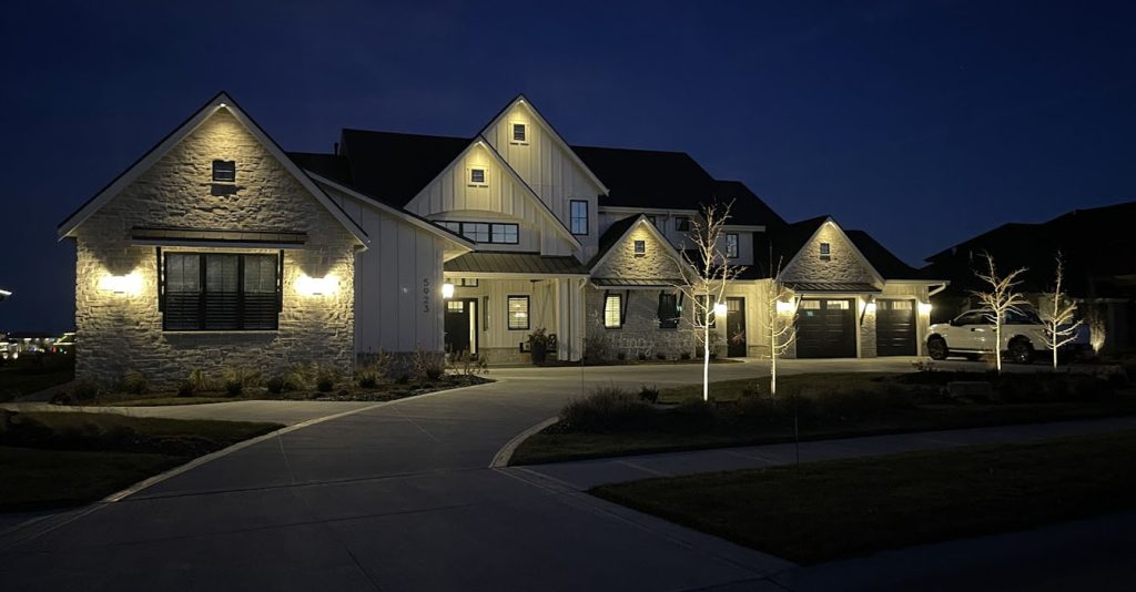 White farmhouse-style home features warm architectural uplighting on multiple stone and siding peaks. Modern sconces illuminate the garage and windows, while ground-level fixtures highlight young trees along the driveway.