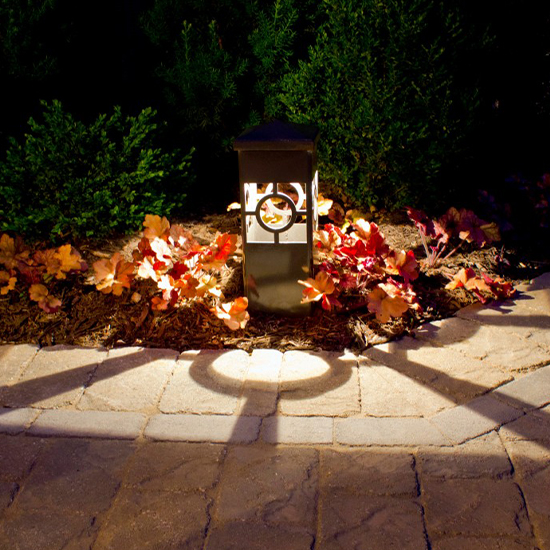 A decorative path light with a circular cutout sits within a garden bed, illuminating red and green foliage. It casts a distinct, ring-shaped shadow across the textured stone pavers of a walkway.