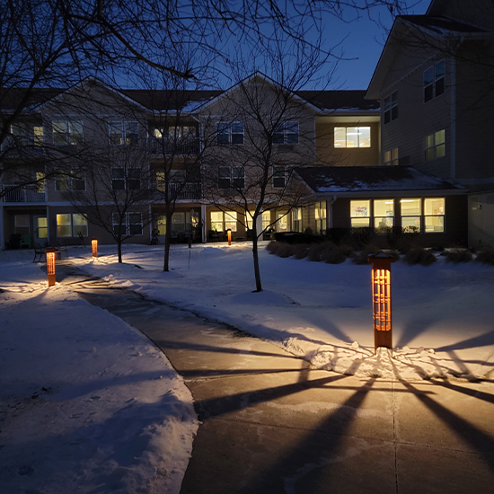 Bollard path lights with a distinctive wood-slat design line a winding sidewalk through a snowy courtyard. These lights cast long, dramatic shadows across the snow and pavement toward a multi-story residential building.