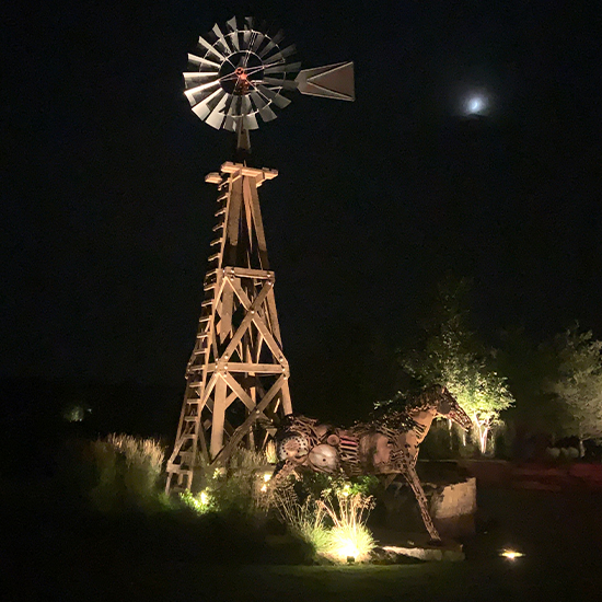 A metal windmill and horse sculpture stand prominently in this dark outdoor landscape. Bright ground spotlights illuminate the structures from below, casting a warm glow against the backdrop of a faint, distant moon.