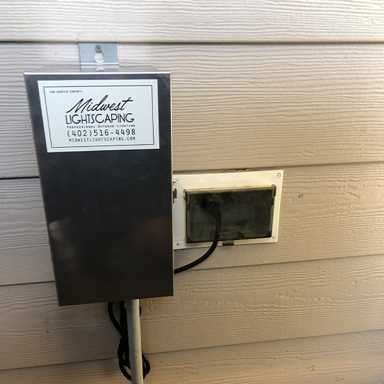 Metallic control box featuring "Midwest Lightscaping" contact information is mounted on a tan-sided wall. A black electrical cord connects the box to an adjacent covered outdoor outlet for professional power management.