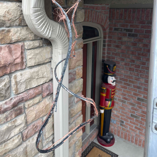 Damaged electrical wiring with frayed insulation and exposed copper strands hangs precariously from a tan gutter downspout. Background elements include a stone facade and a decorative toy soldier nutcracker standing near a brick entryway.