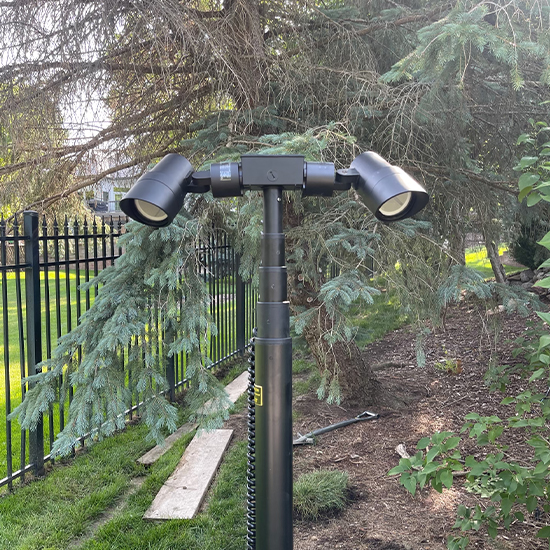 Dual black spotlights are mounted atop a telescoping pole, positioned within a landscaped yard near a black metal fence. Surrounding evergreen trees and mulch indicate a professional outdoor lighting installation for property enhancement.