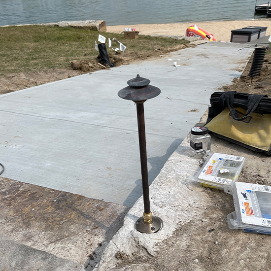 Tiered copper path light stands securely installed within a stone border adjacent to a fresh concrete walkway. Nearby tools and hardware indicate an active professional installation project situated by a lakefront property.