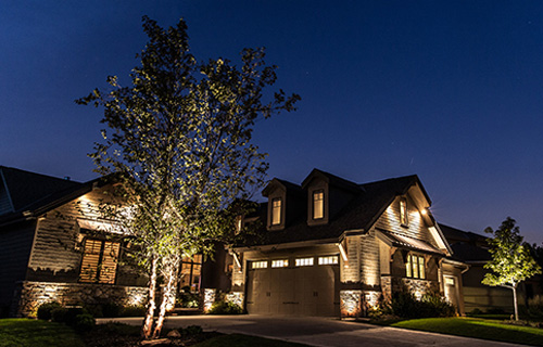 This contemporary two-story home features a striking combination of stone masonry and horizontal siding under a clear evening sky. Strategic warm uplighting illuminates a multi-trunked tree in the foreground and the home's textured architectural details.