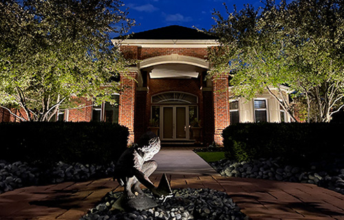 This residential scene features a red brick home with a central arched entryway at night. In the foreground, a sculpture of a child playing with a boat is brightly illuminated. Warm uplighting highlights the surrounding mature trees and the architectural columns of the house.