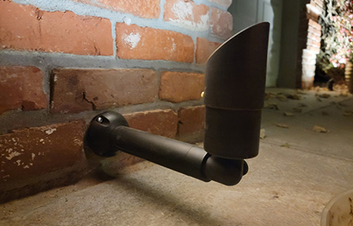 This outdoor landscape fixture is a dark, cylindrical directional spotlight mounted to a red brick wall. It features a long extension arm and an angled shroud designed to aim light upward or toward specific architectural features.