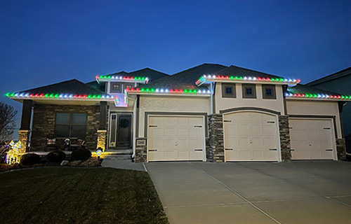 Vibrant green, red and white holiday string lights trace the roofline gables of this dark brick residence. Symmetrical warm spotlights illuminate the lower facade, casting vertical glows that accentuate the home's textured masonry at night.