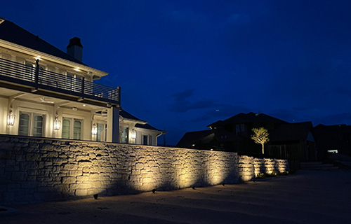 A light-colored stone wall is dramatically illuminated by ground-mounted spotlights at night. Warm lights on the adjacent multi-story house highlight architectural details under a deep blue sky.