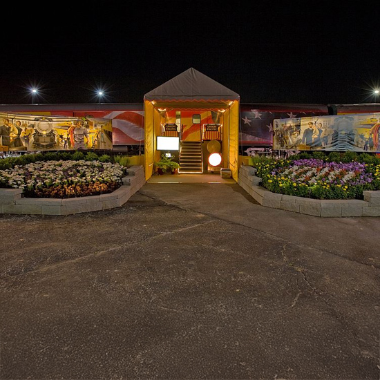 A brightly lit yellow tent serves as an entryway to a train car decorated with American flag motifs and murals. Elevated flower beds on either side of the asphalt path are subtly illuminated by integrated landscape lighting.