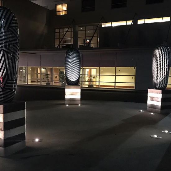 Several large, patterned sculptures are illuminated from below by small well lights embedded in the surrounding pavement.