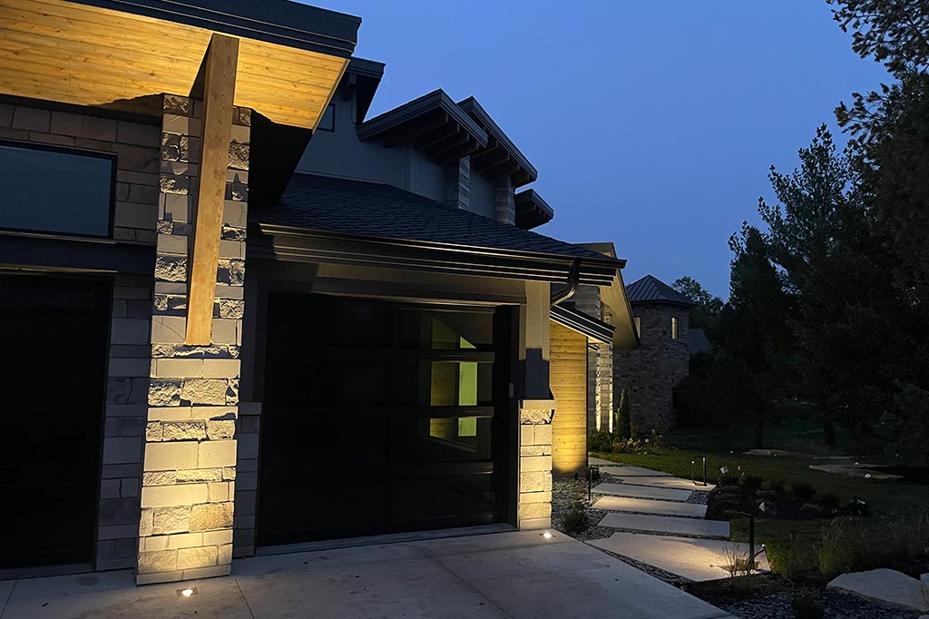 Modern home features warm accent lighting on stone pillars and wooden eaves above a glass garage door. Minimalist path lights and recessed well lights guide the way along a concrete walkway into the distance in Douglas County.