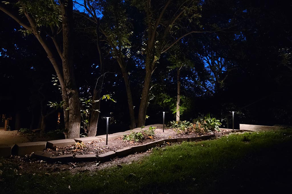 Low stone border encircles a raised garden bed at the edge of a wooded area. Modern, slender path lights stand within the mulch to illuminate the plants and nearby tree trunks with a cool white glow in Douglas County.