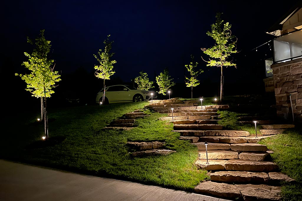 Stone stairs leading up a grassy hill are illuminated by multiple slender path lights. Warm uplighting highlights a row of young ornamental trees along the incline, while a white car is parked at the top near the home.