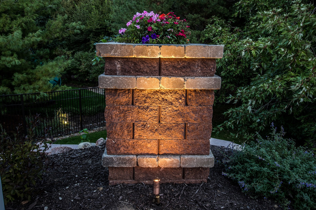 Stone pillar topped with colorful flowers features integrated hardscape lights tucked under the ledge. A small ground-mounted accent light at the base provides additional upward illumination to showcase the brick texture in Douglas County.