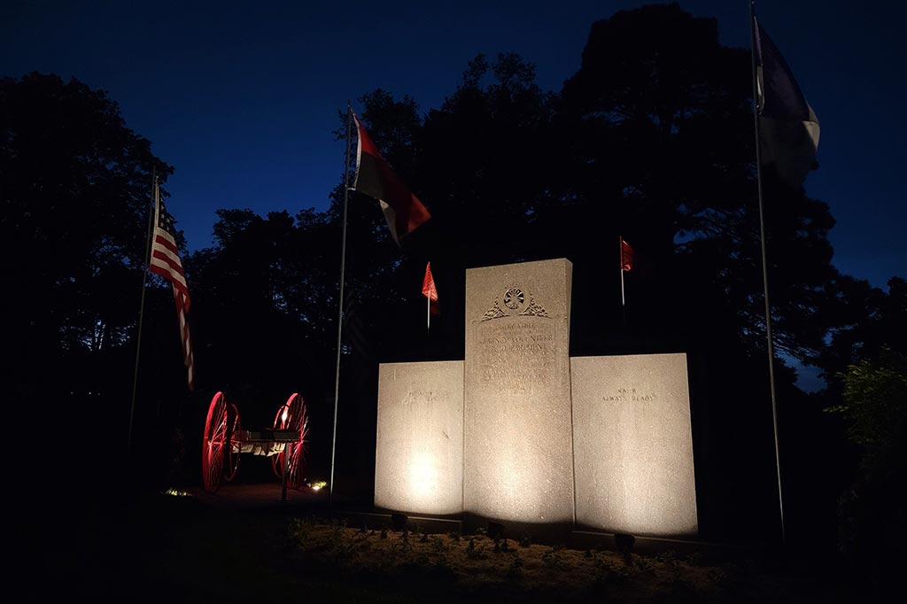 Stone memorial wall stands at the center of a dark landscape flanked by several flagpoles. Ground-mounted uplighting creates a bright wash across the granite surfaces, while smaller accent lights highlight the wheels of a nearby vintage cart in Douglas County.