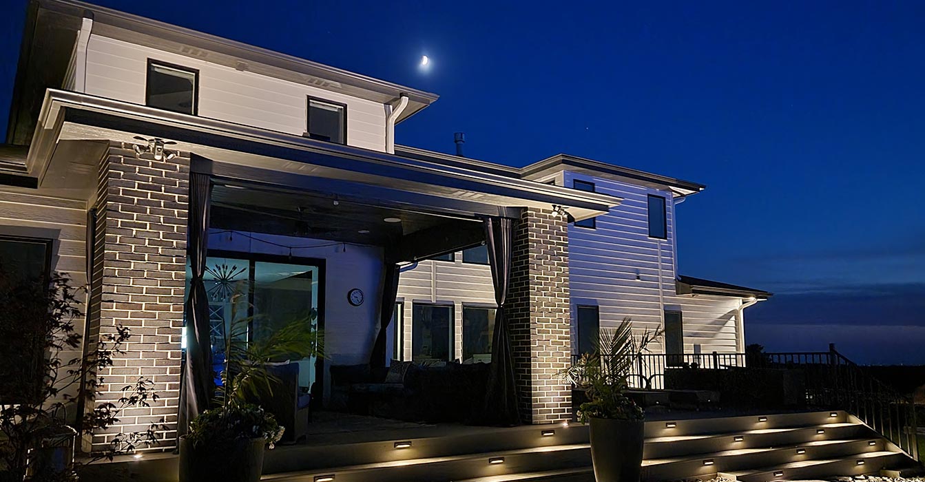 This two-story white farmhouse features a covered patio area with prominent brick pillars. The outdoor space utilizes several lighting techniques to enhance safety and aesthetics.