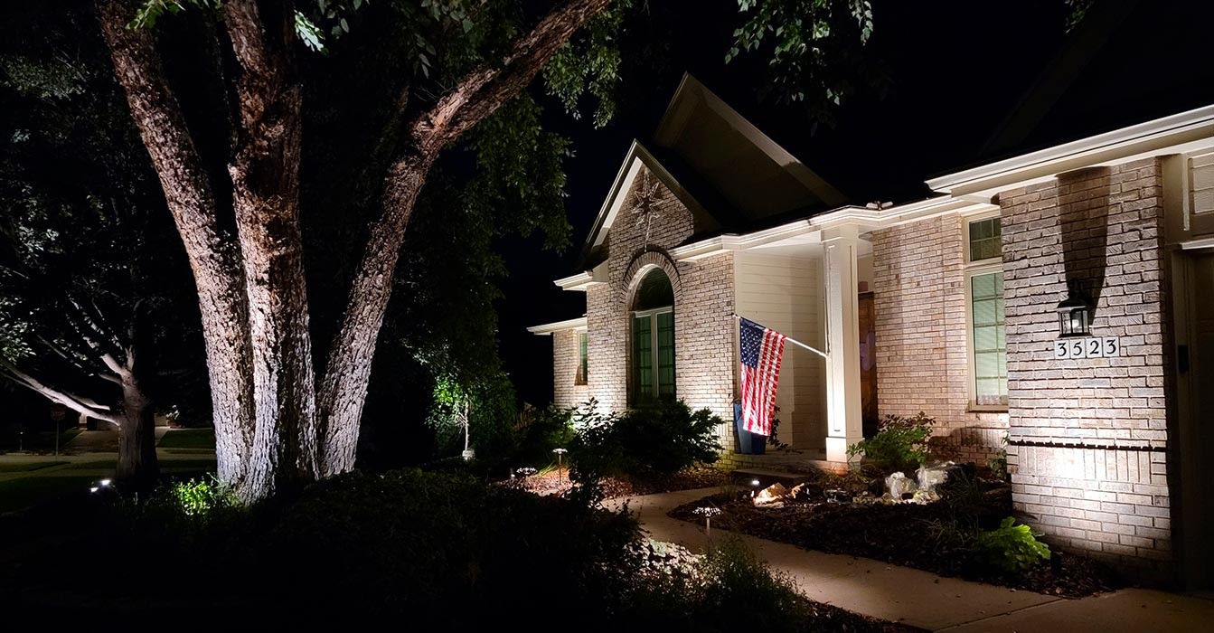 Brick exterior of a ranch-style home is highlighted by several uplights that create a warm wash across the stone facade and architectural peaks.