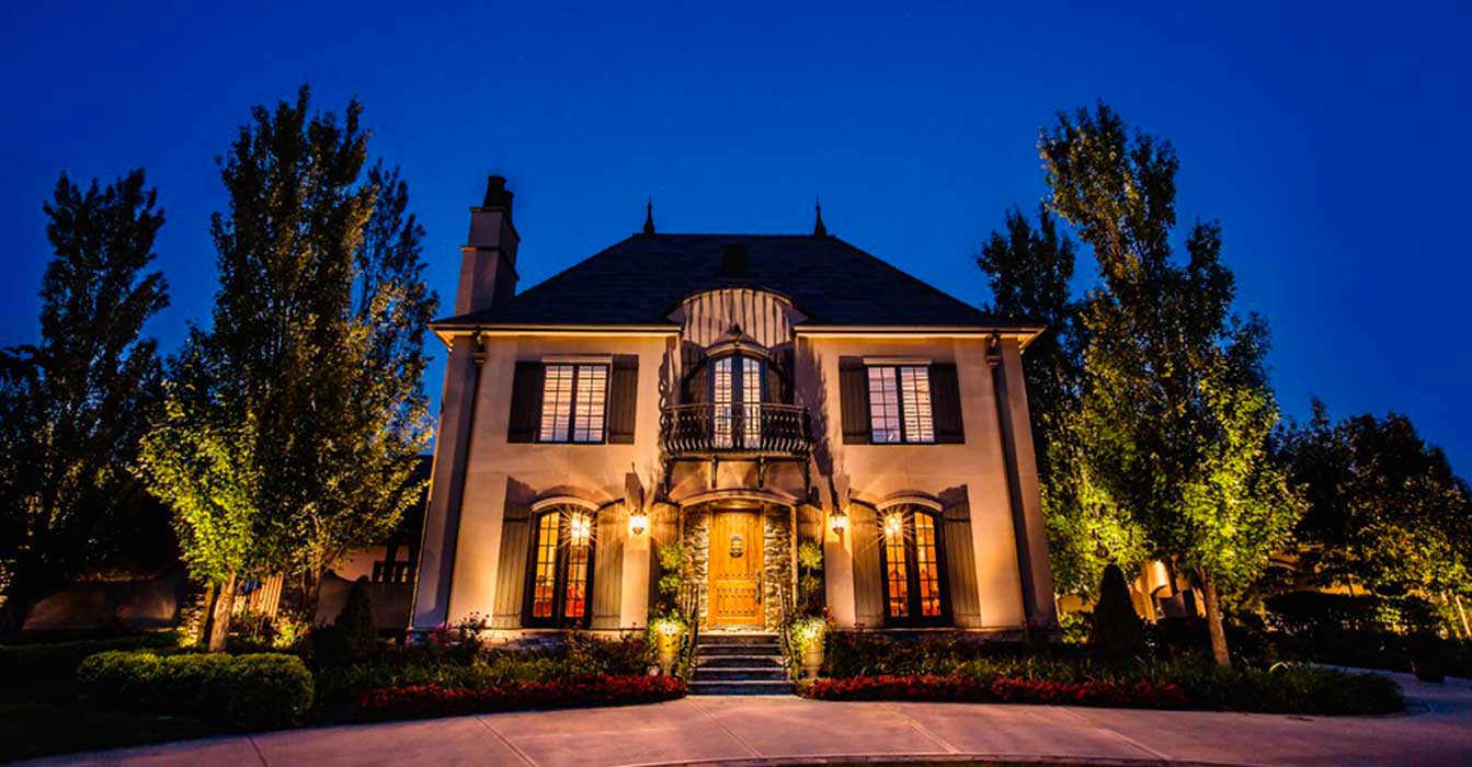 This two-story French country home features a steep roof, stone accents, and a central balcony. Ground fixtures use uplighting to create a warm light wash on the facade and tall flanking trees.