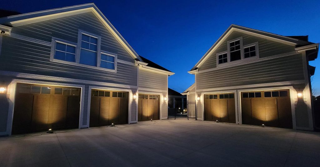 This light-colored home features multiple gables and a large concrete driveway. Ground-level fixtures provide uplighting to create a warm wash on five dark garage doors.