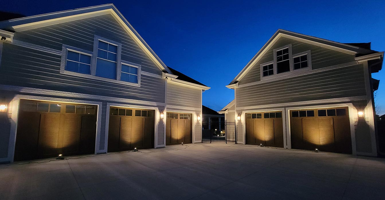 This light-colored home features multiple gables and a large concrete driveway. Ground-level fixtures provide uplighting to create a warm wash on five dark garage doors.