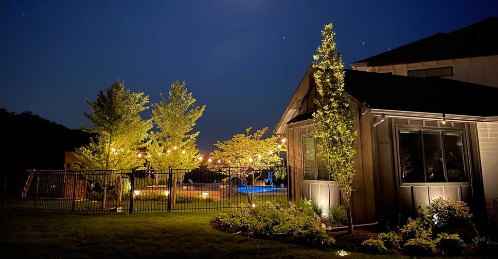 Grey board-and-batten siding covers this home. Ground uplighting accents the trees, and string lights add a warm glow. This creates a bright wash for night use.