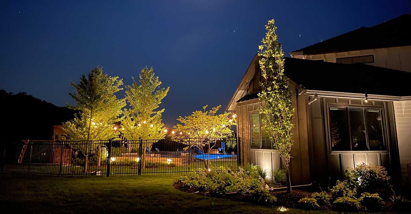 Grey board-and-batten siding covers this home. Ground uplighting accents the trees, and string lights add a warm glow. This creates a bright wash for night use.