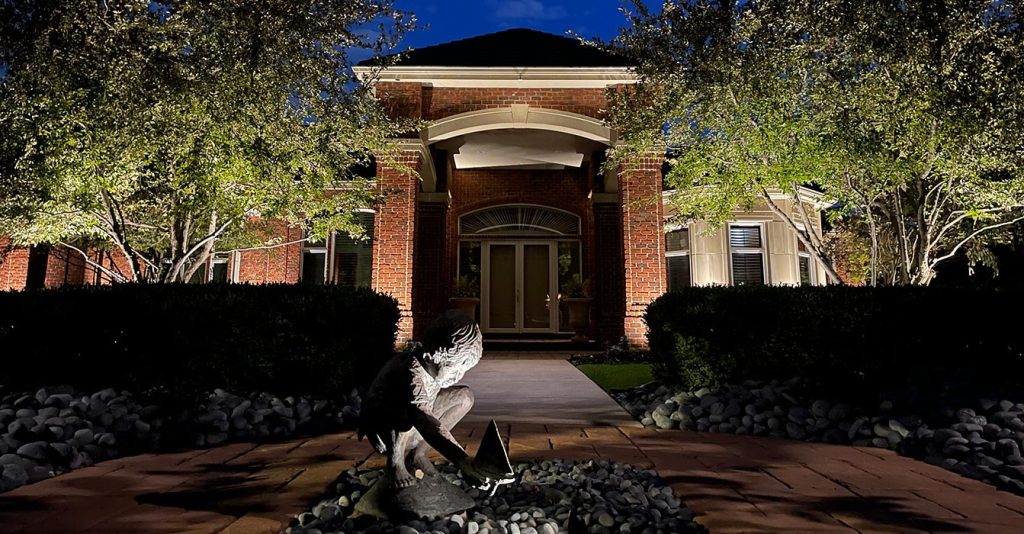 A nighttime view of a grand brick home featuring uplit pillars and trees, with a central spotlight highlighting a sculpture of a child with a sailboat in the foreground.
