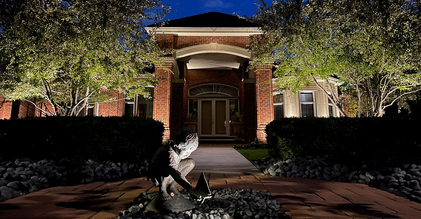 A nighttime view of a grand brick home featuring uplit pillars and trees, with a central spotlight highlighting a sculpture of a child with a sailboat in the foreground.