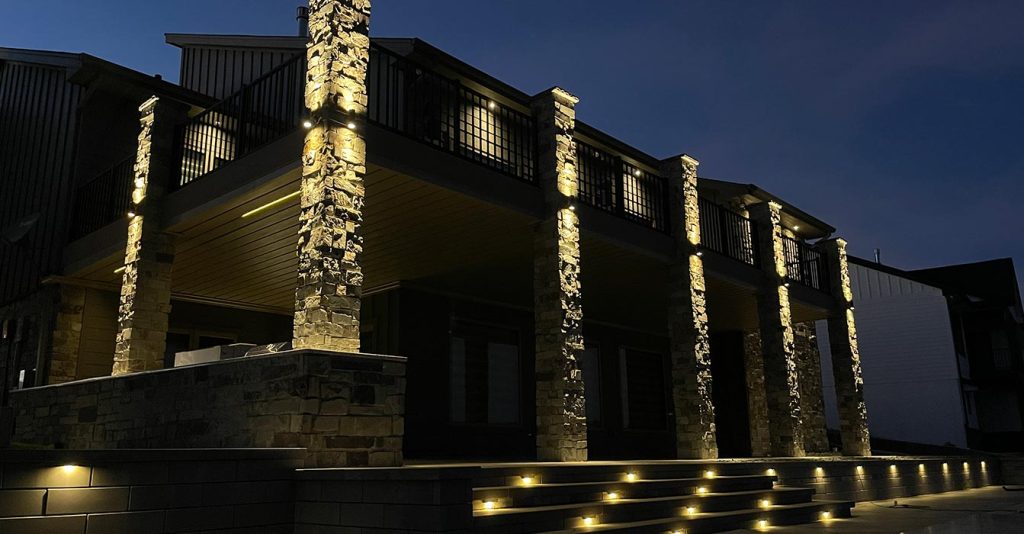 A nighttime view of a modern house featuring dramatically uplit stone pillars, a tiered stone staircase with integrated path lights, and recessed accent lighting on stone retaining walls.