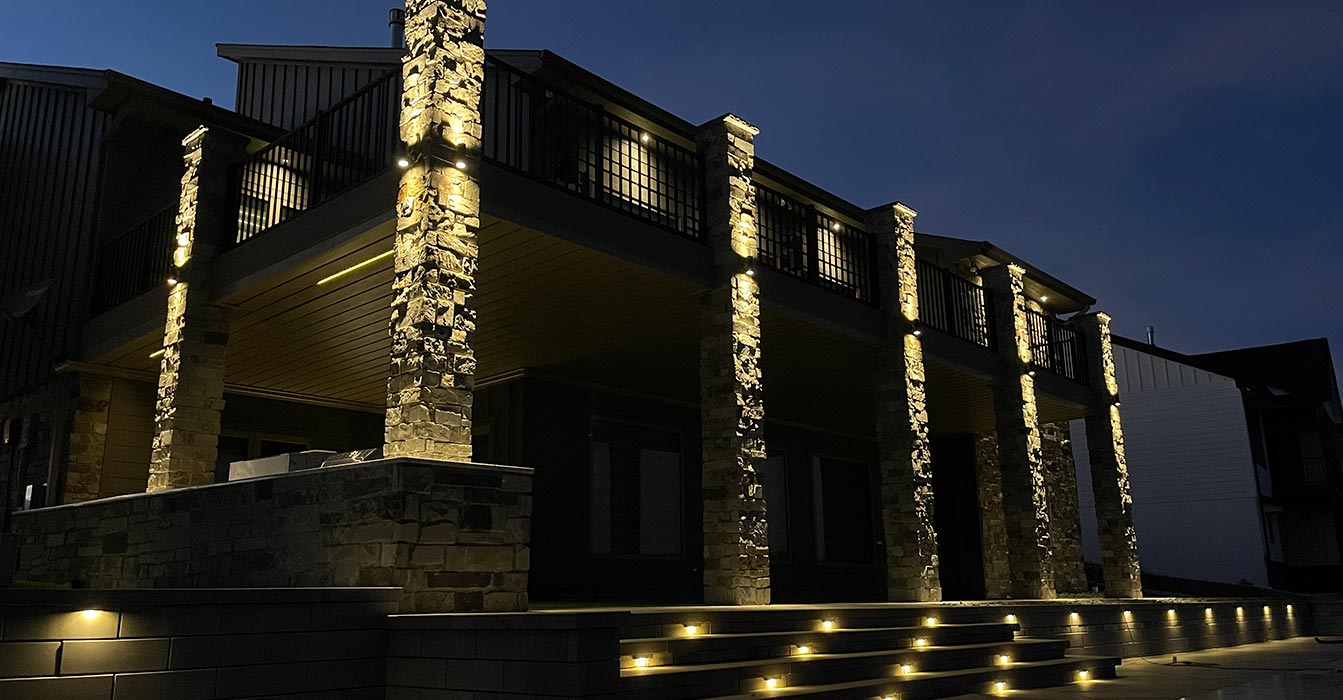 A nighttime view of a modern house featuring dramatically uplit stone pillars, a tiered stone staircase with integrated path lights, and recessed accent lighting on stone retaining walls.
