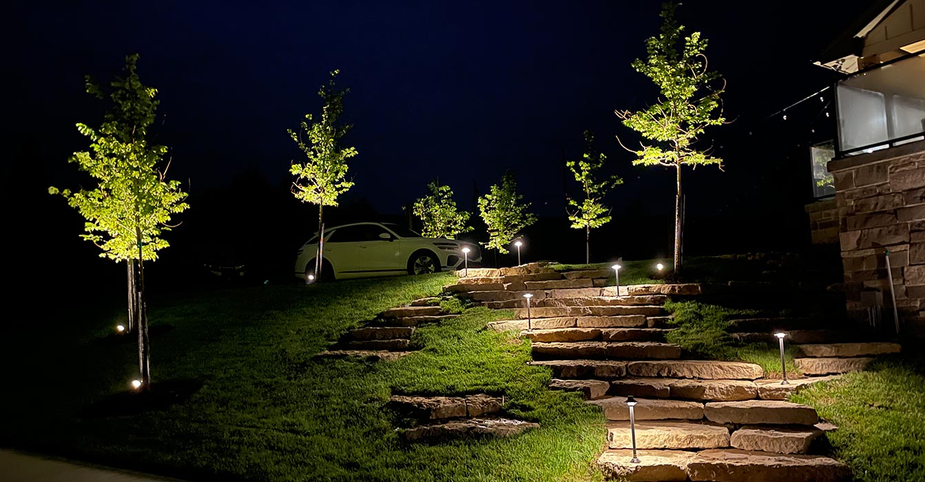 A nighttime view of a modern house featuring dramatically uplit stone pillars, a tiered stone staircase with integrated path lights, and recessed accent lighting on retaining walls.