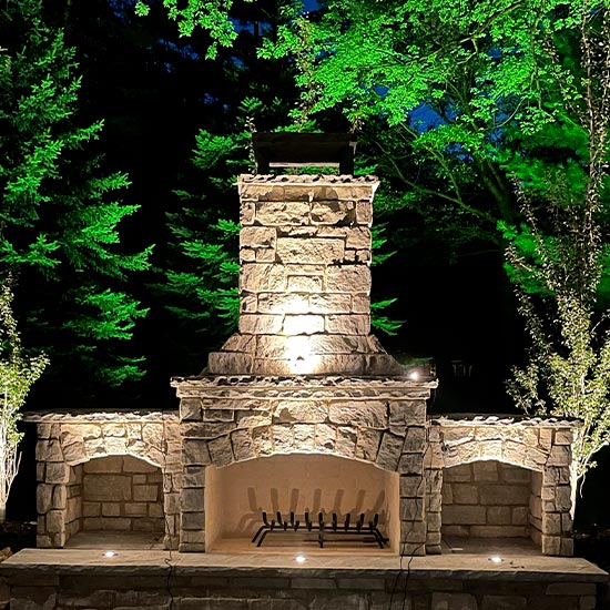 A nighttime view of a gabled house featuring uplit stone pillars, illuminated tiered stone retaining walls with recessed sconces, and a multi-level stone staircase guided by path lights.