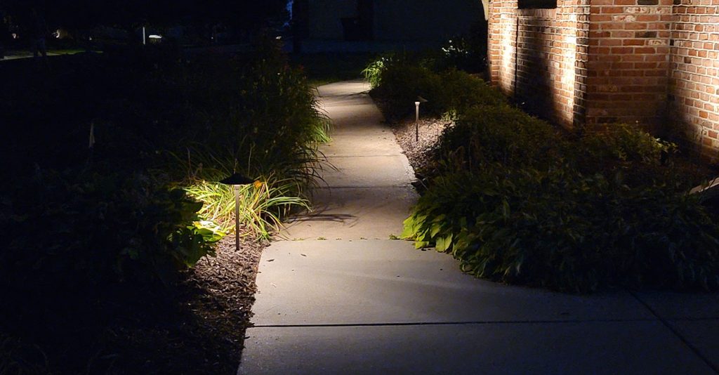 A concrete garden walkway at night illuminated by small, warm path lights, with architectural uplighting on a brick wall and light focusing on low-lying shrubs and hostas along the path.