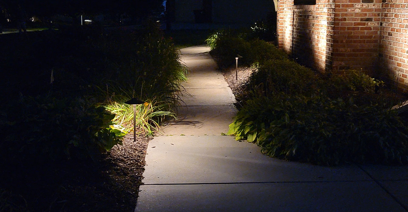 A concrete garden walkway at night illuminated by small, warm path lights, with architectural uplighting on a brick wall and light focusing on low-lying shrubs and hostas along the path.