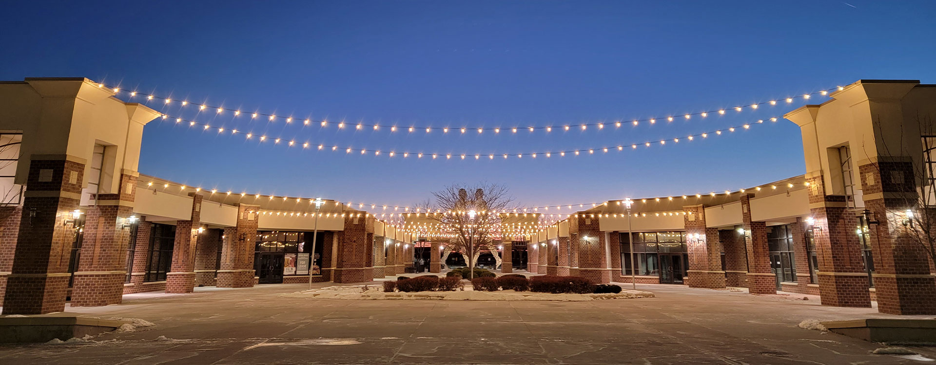 A wide-angle view of an outdoor brick shopping plaza at dusk, featuring multiple strings of warm globe lights crisscrossing above a central concrete courtyard.