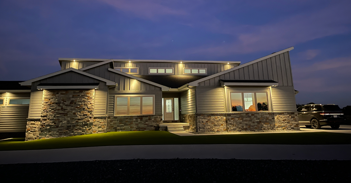 A modern house with grey vertical siding and stone accents at night, featuring architectural downlighting along the roofline and stone facade, with a dark SUV parked in the driveway.