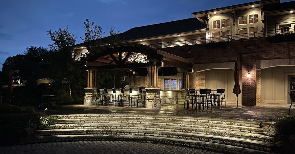 A stone patio at night featuring a large wooden pergola with an illuminated chandelier over a built-in bar, with curved stone steps and architectural lighting on the brick building in the background.